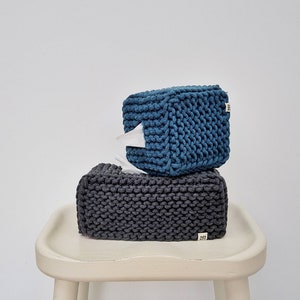 May include: Two knitted tissue box covers, one dark grey and one teal blue, stacked on a white stool.