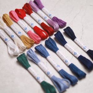 May include: A collection of colorful embroidery floss in various shades, including white, gold, pink, red, blue, green, and purple. Each skein is neatly wrapped with a paper label featuring Japanese text and a floral design. Ideal for embroidery and craft projects.