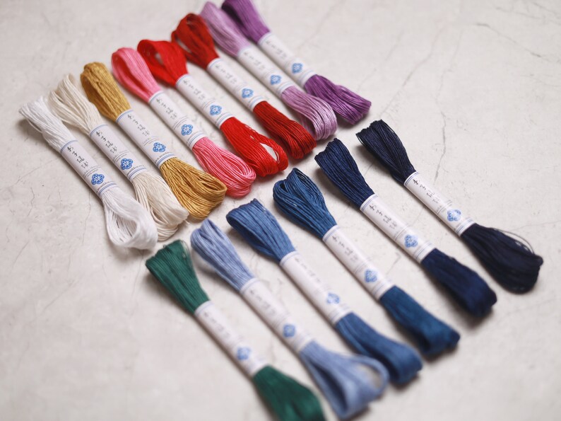 May include: A collection of colorful embroidery floss in various shades, including white, gold, pink, red, blue, green, and purple. Each skein is neatly wrapped with a paper label featuring Japanese text and a floral design. Ideal for embroidery and craft projects.