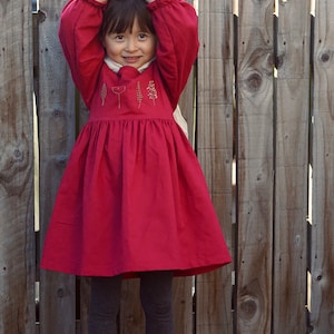 Puede incluir: Una ni&ntilde;a peque&ntilde;a con un vestido rojo con detalles florales bordados, de pie frente a una valla de madera. Est&aacute; sonriendo y tiene las manos juntas frente a su cara.