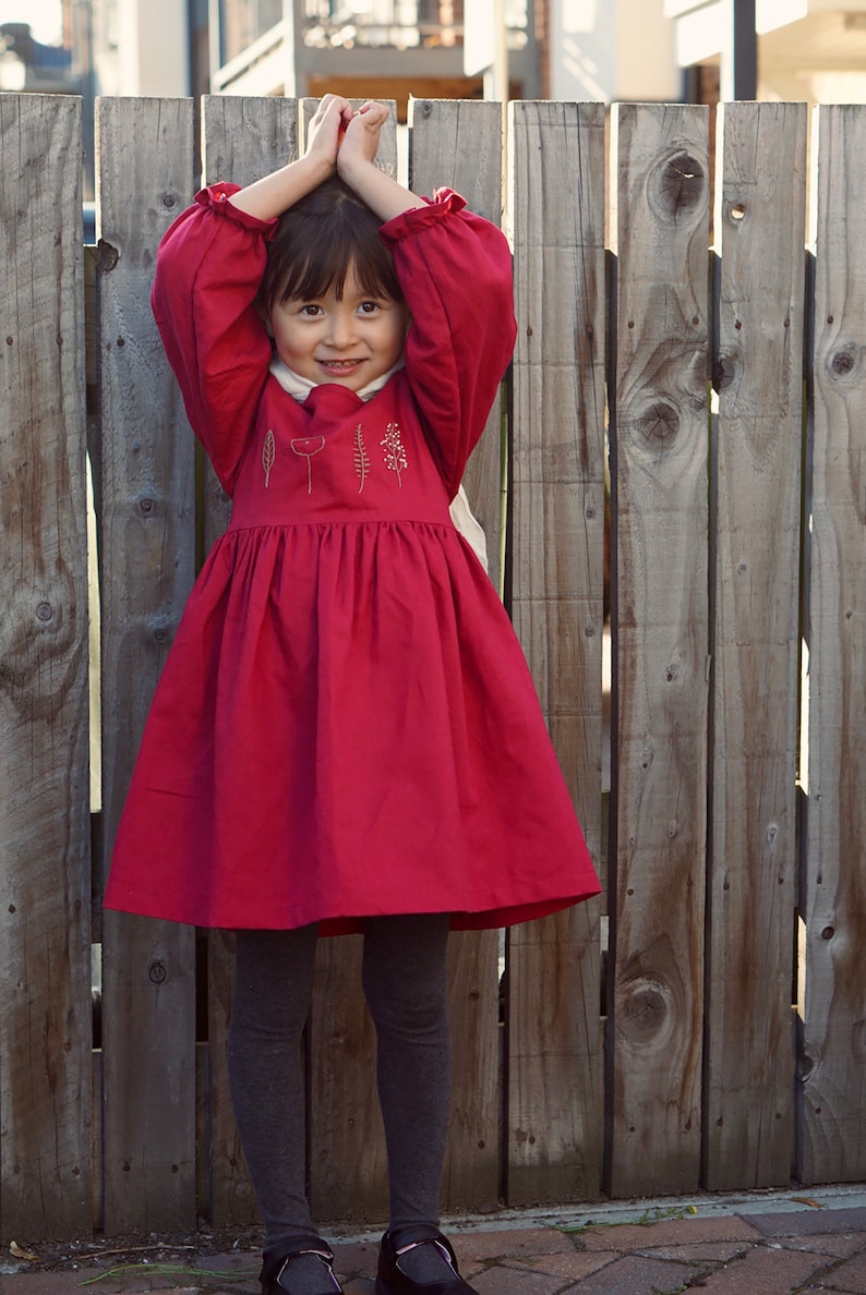 Puede incluir: Una ni&ntilde;a peque&ntilde;a con un vestido rojo con detalles florales bordados, de pie frente a una valla de madera. Est&aacute; sonriendo y tiene las manos juntas frente a su cara.