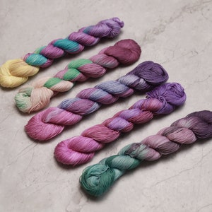 May include: Six skeins of yarn in various shades of pink, purple, and green. The yarn is arranged in a row, with the lightest shade at the top and the darkest shade at the bottom. visible creative mending thread