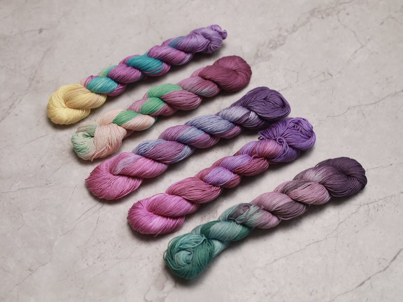 May include: Six skeins of yarn in various shades of pink, purple, and green. The yarn is arranged in a row, with the lightest shade at the top and the darkest shade at the bottom. visible creative mending thread