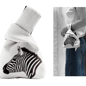 Pu&ograve; includere: Borsa tote in tela bianca con un design a testa di zebra nera. La borsa &egrave; piegata e legata in cima.