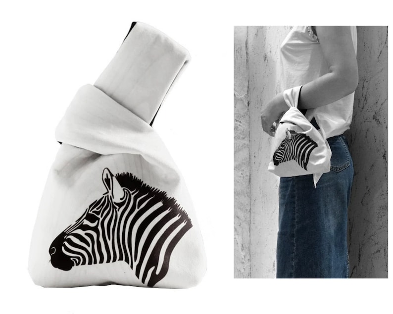 Pu&ograve; includere: Borsa tote in tela bianca con un design a testa di zebra nera. La borsa &egrave; piegata e legata in cima.