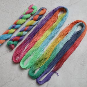 May include: Two twisted hanks of multi-colored yarn featuring pink, green, orange, and blue hues, alongside two long, loose loops of yarn in a gradient of pink, orange, yellow, light blue, green, dark blue, and purple strands.