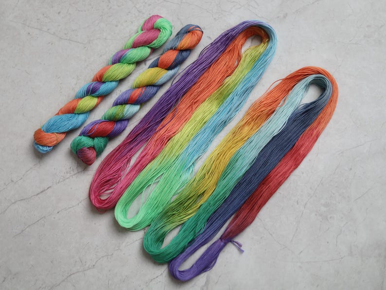 May include: Two twisted hanks of multi-colored yarn featuring pink, green, orange, and blue hues, alongside two long, loose loops of yarn in a gradient of pink, orange, yellow, light blue, green, dark blue, and purple strands.