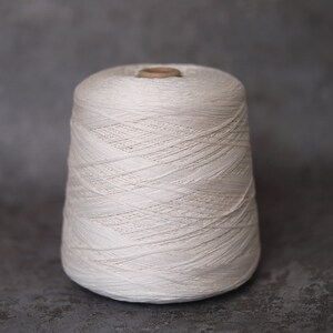 May include: A large spool of off-white yarn, tightly wound and cone-shaped. The yarn appears to be a natural fiber, with visible texture and subtle variations in color. The spool sits against a neutral gray background, highlighting the yarn's texture.