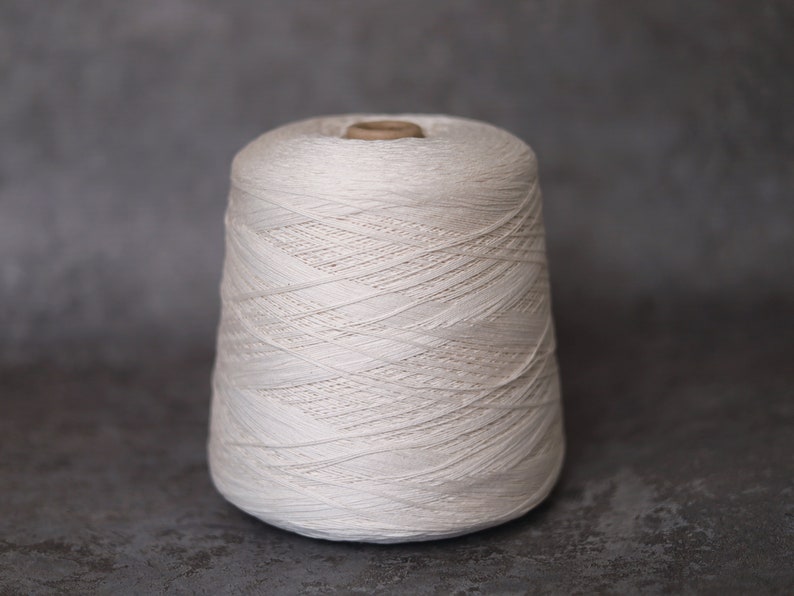 May include: A large spool of off-white yarn, tightly wound and cone-shaped. The yarn appears to be a natural fiber, with visible texture and subtle variations in color. The spool sits against a neutral gray background, highlighting the yarn's texture.