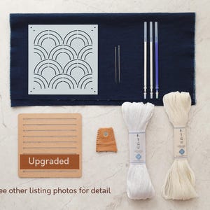 Könnte beinhalten: Ein Bastelset zum Sticken mit einem blauen Stoff, einer weißen Schablone mit geometrischem Muster, zwei Spulen weißen Stickgarns, drei Sticknadeln, einem Fingerhut und einem Holzrahmen mit dem aufgedruckten Wort "Upgraded".