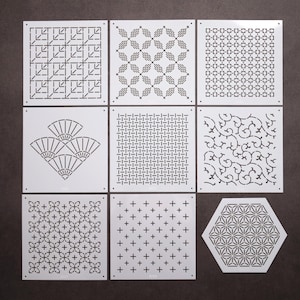 Può includere: Una collezione di nove stencil bianchi quadrati ed esagonali con vari motivi geometrici, tra cui forme a ventaglio, croci, texture intrecciate e complessi disegni floreali, ciascuno dotato di piccoli fori negli angoli per l'allineamento.