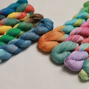 Op de afbeelding: Een verzameling kleurrijke garenklossen in verschillende tinten blauw, groen, geel, oranje, roze en paars. Het garen is in twee groepen gerangschikt, met de ene groep bovenop de andere.