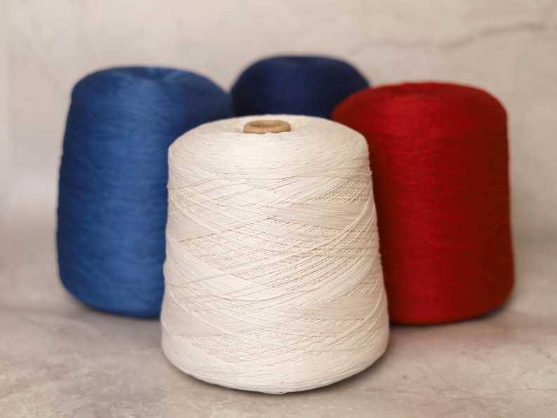 May include: Four spools of yarn in shades of blue, red, and white. The yarn is tightly wound on cardboard cores. The white spool is in the foreground, with the other spools arranged behind it. The yarn has a smooth texture.