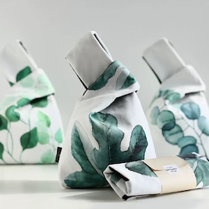 Pu&ograve; includere: Tre sacchetti di tessuto bianco con motivi di foglie verdi. I sacchetti sono legati in cima con un nodo. I sacchetti sono su una superficie bianca. Il sacchetto al centro ha un'etichetta che dice "botanical goods".