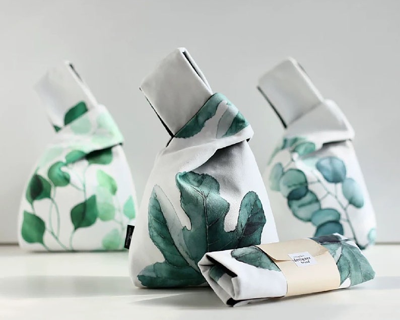 Pu&ograve; includere: Tre sacchetti di tessuto bianco con motivi di foglie verdi. I sacchetti sono legati in cima con un nodo. I sacchetti sono su una superficie bianca. Il sacchetto al centro ha un'etichetta che dice "botanical goods".