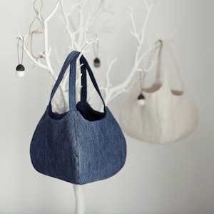 May include: Two fabric tote bags hanging on a white branch. The bag on the left is a dark blue denim tote bag. The bag on the right is a light beige tote bag.