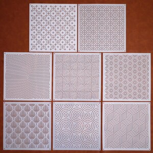 May include: Nine white square stencils featuring various geometric patterns. The designs include circles, lines, and squares.