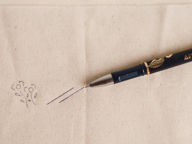 May include: A black pen with a gold wing design on the barrel is lying on a beige fabric surface. The pen is pointing to the left and has drawn a small flower and two parallel lines.