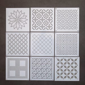 Peut inclure: Neuf pochoirs carrés blancs avec différents motifs géométriques, notamment une fleur, des diamants, des carrés et un motif de feuilles.