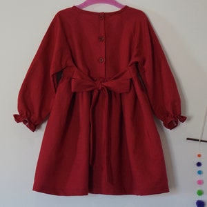 Puede incluir: Un vestido rojo de manga larga con un lazo en la cintura y cierre de botones en la espalda.