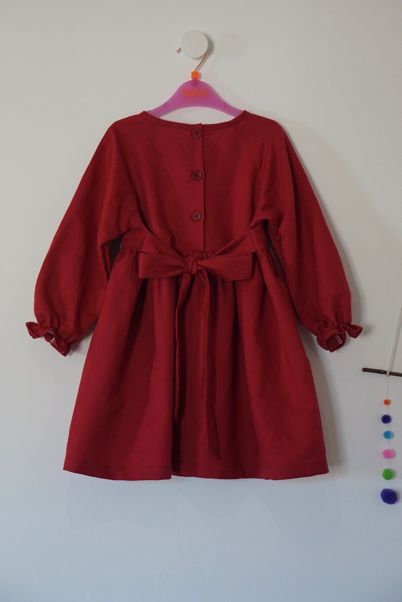 Puede incluir: Un vestido rojo de manga larga con un lazo en la cintura y cierre de botones en la espalda.