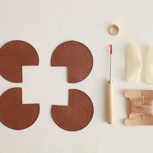 May include: Four brown leather pieces with cutouts, a spool of brown thread, a small gold ring, two white thimbles, and a small wooden tool with a red tip.