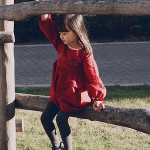 Puede incluir: Una ni&ntilde;a peque&ntilde;a con un vestido rojo con detalles bordados se sienta en un tronco de madera. Lleva medias negras y zapatos negros.