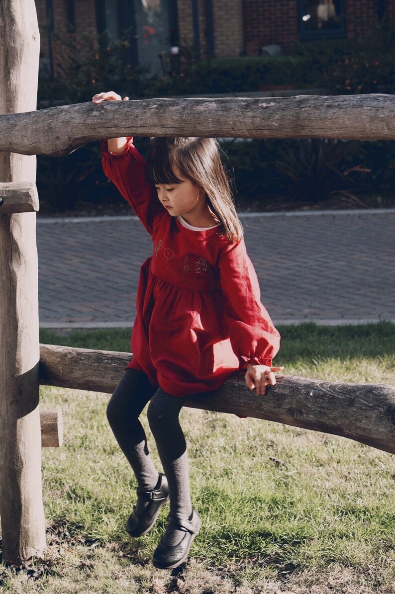 Puede incluir: Una ni&ntilde;a peque&ntilde;a con un vestido rojo con detalles bordados se sienta en un tronco de madera. Lleva medias negras y zapatos negros.