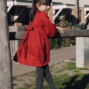 Puede incluir: Una ni&ntilde;a peque&ntilde;a con un vestido rojo con un lazo en la espalda, medias negras y zapatos negros. Est&aacute; de pie sobre una valla de madera y mirando por encima del hombro.
