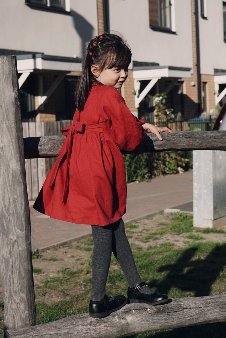 Puede incluir: Una ni&ntilde;a peque&ntilde;a con un vestido rojo con un lazo en la espalda, medias negras y zapatos negros. Est&aacute; de pie sobre una valla de madera y mirando por encima del hombro.