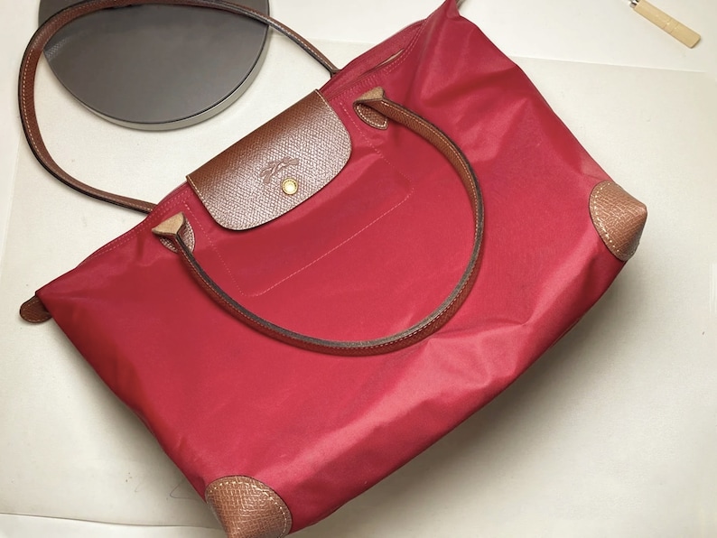Puede incluir: Un bolso tote de nylon rojo con ribetes de cuero marr&oacute;n y un tirador de cremallera dorado. El bolso tiene un asa superior &uacute;nica y dos correas para el hombro.