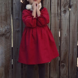 Puede incluir: Una ni&ntilde;a peque&ntilde;a con un vestido rojo, una bufanda blanca y zapatos negros se para frente a una valla de madera.