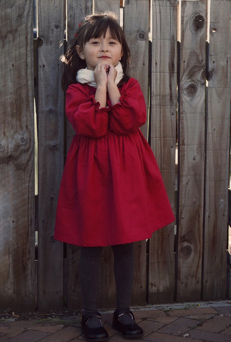 Puede incluir: Una ni&ntilde;a peque&ntilde;a con un vestido rojo, una bufanda blanca y zapatos negros se para frente a una valla de madera.