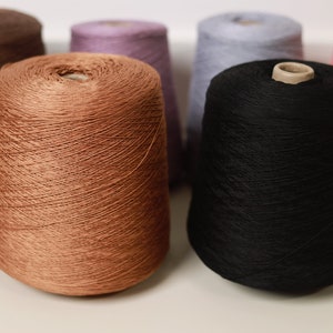 Könnte beinhalten: Sechs Garnrollen in verschiedenen Farben, darunter Braun, Rosa, Hellblau und ein dunkleres Braun. Die Rollen sind in einer Reihe auf einer weißen Oberfläche angeordnet.