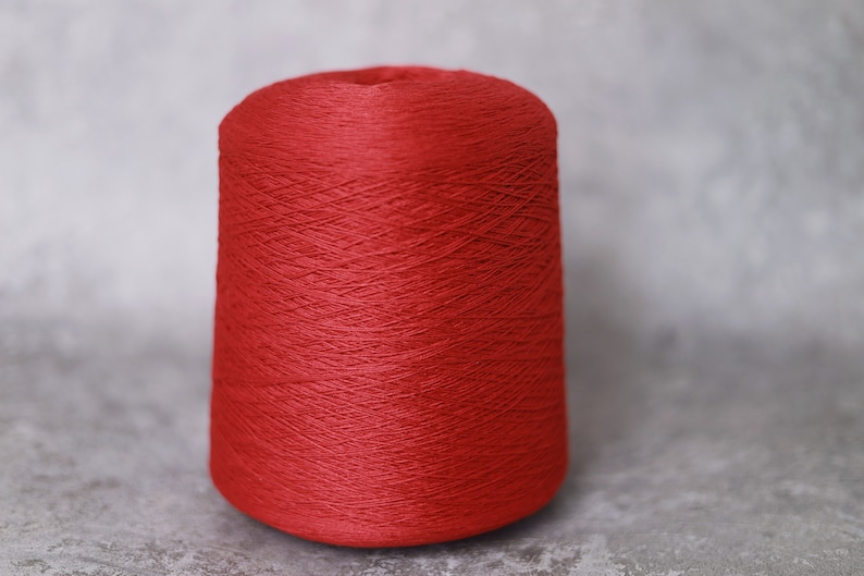 May include: A spool of vibrant red yarn, tightly wound and cone-shaped. The yarn has a smooth texture and is suitable for various knitting and crochet projects. The background is a neutral gray.