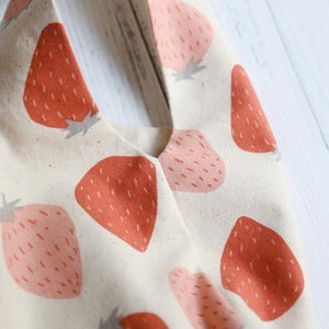 Pu&ograve; includere: Una borsa tote in tela bianca con una stampa di fragole. Le fragole sono rosse e rosa su uno sfondo bianco.