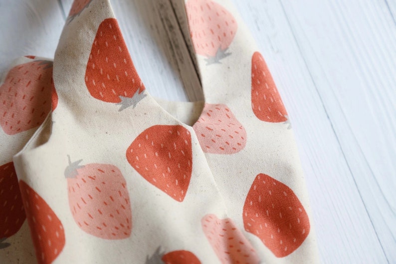 Pu&ograve; includere: Una borsa tote in tela bianca con una stampa di fragole. Le fragole sono rosse e rosa su uno sfondo bianco.