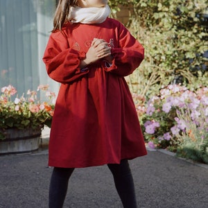 Puede incluir: Una ni&ntilde;a peque&ntilde;a con un vestido rojo con detalles bordados, medias negras y zapatos negros. Est&aacute; de pie frente a una pared de ladrillo y un jard&iacute;n de flores.