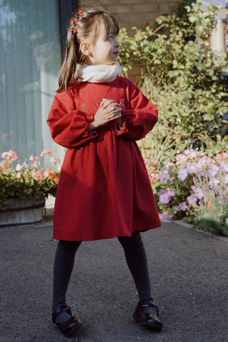 Puede incluir: Una ni&ntilde;a peque&ntilde;a con un vestido rojo con detalles bordados, medias negras y zapatos negros. Est&aacute; de pie frente a una pared de ladrillo y un jard&iacute;n de flores.