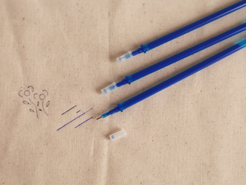 May include: Three blue ink pens with clear plastic caps on a beige fabric surface.