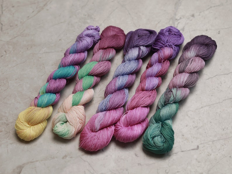 May include: Five skeins of hand-dyed yarn in various shades of pink, purple, green, and yellow. The yarn is twisted and has a subtle ombre effect. visible creative mending quilting thread