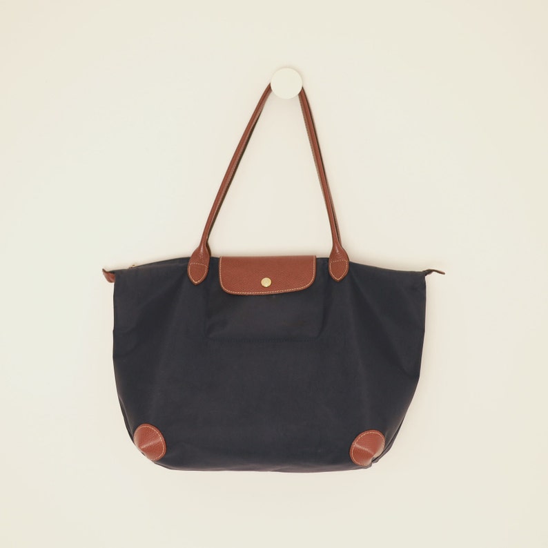 Puede incluir: Un bolso tote negro con detalles en cuero marr&oacute;n y un peque&ntilde;o bolsillo en la parte delantera. El bolso est&aacute; colgado de un gancho blanco.