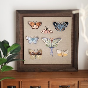May include: A square dark wood frame containing a beige fabric backing with seven embroidered insects. The collection includes various butterflies and moths in shades of orange, blue, brown, pink, green, and yellow, arranged on the fabric surface.