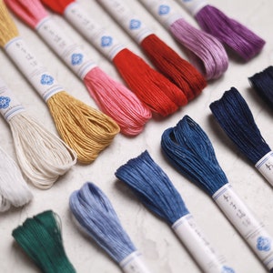 May include: A collection of colorful embroidery floss skeins in various shades, including white, pink, red, gold, blue, and purple. Each skein is wrapped with a paper label featuring Japanese text and a blue floral design.