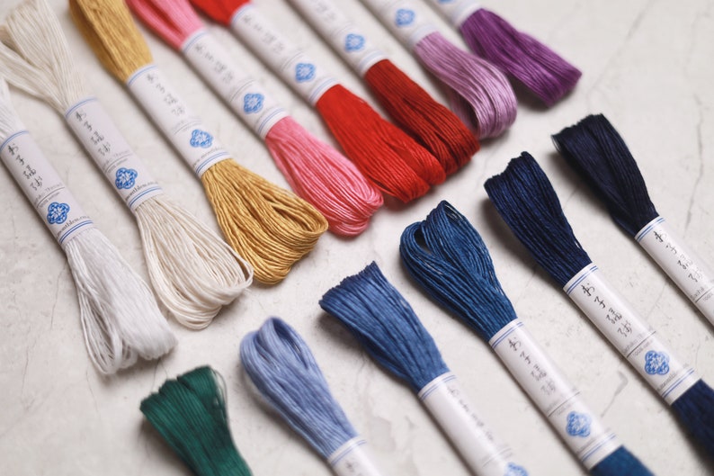 May include: A collection of colorful embroidery floss skeins in various shades, including white, pink, red, gold, blue, and purple. Each skein is wrapped with a paper label featuring Japanese text and a blue floral design.