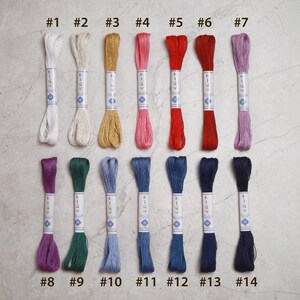 May include: A collection of fourteen skeins of embroidery floss in various colors, including white, beige, gold, pink, red, and purple. Each skein is labeled with a number from 1 to 14. The floss is neatly arranged on a light-colored surface.