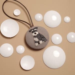 May include: A grey fabric ornament with an embroidered Schnauzer dog design, attached to a brown cord. Several white, round, glossy objects of varying sizes are scattered around the ornament on a beige surface.