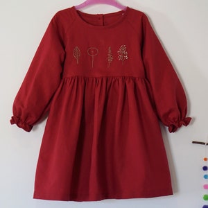 Puede incluir: Un vestido rojo de manga larga con falda fruncida y detalles florales bordados. El vestido tiene un escote redondo y mangas largas con pu&ntilde;os fruncidos.