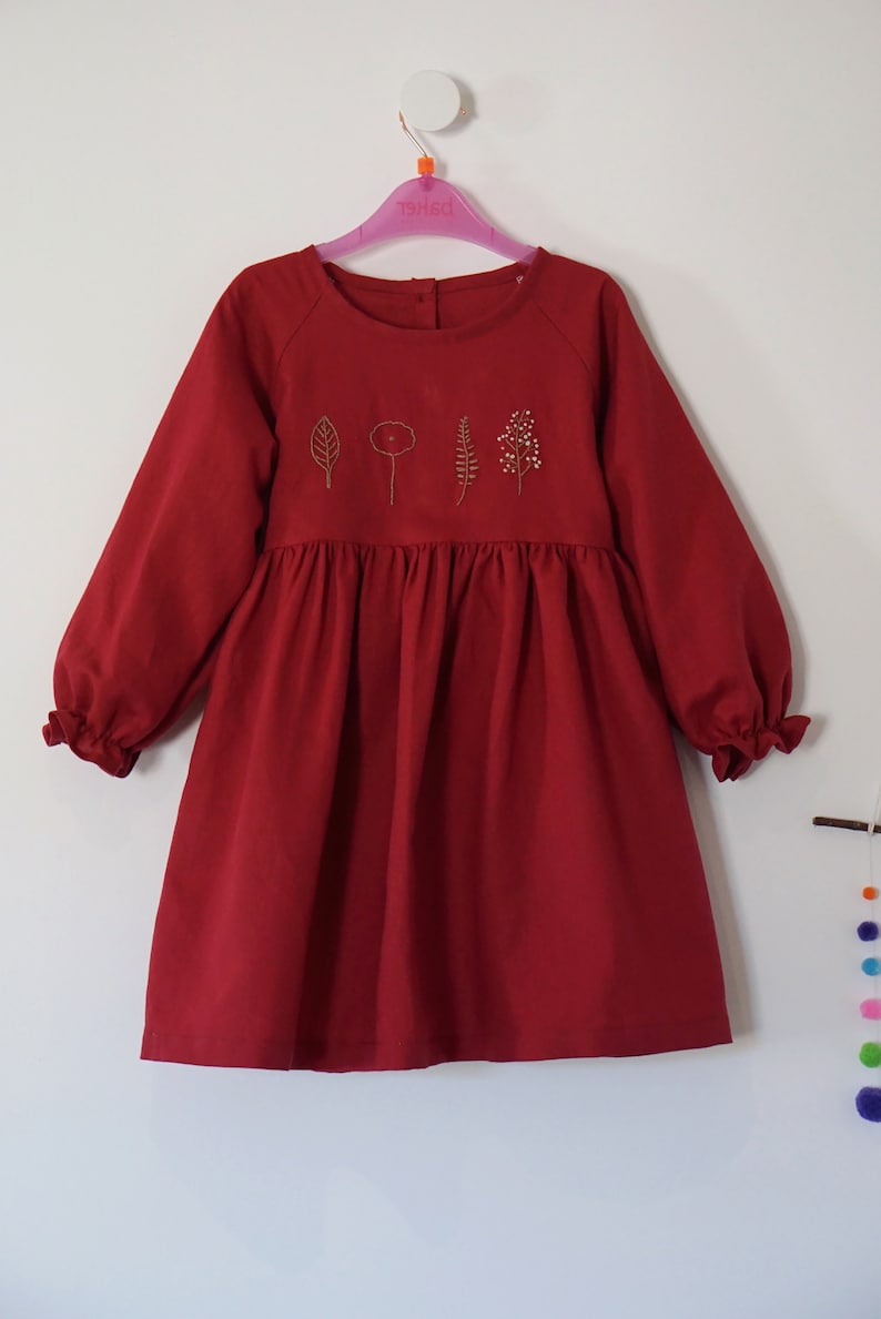 Puede incluir: Un vestido rojo de manga larga con falda fruncida y detalles florales bordados. El vestido tiene un escote redondo y mangas largas con pu&ntilde;os fruncidos.