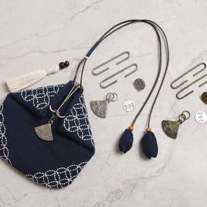 Könnte beinhalten: Marineblaue Stofftasche mit weißem, gesticktem geometrischem Muster, Quaste und langem Kordel. Die Tasche hat einen silbernen Anhänger und wird von Metallverschlüssen, Knöpfen und fächerförmigen Anhängern begleitet. Die Artikel sind auf einer Marmoroberfläche angeordnet.